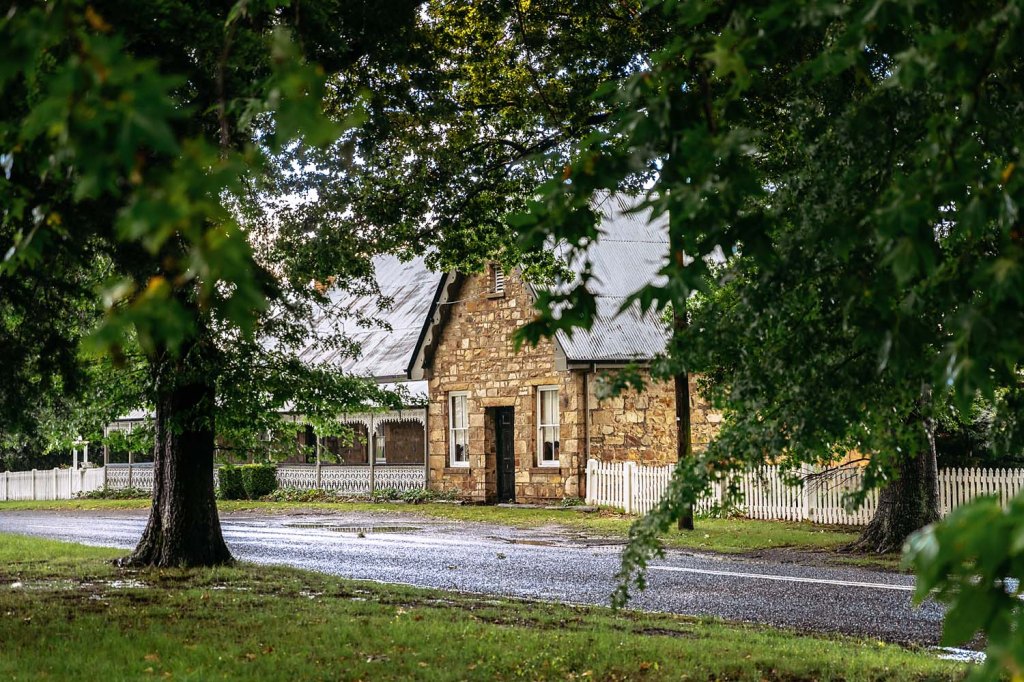 A stone house surrounded by trees and a white picket fence, viewed from a street with wet pavement.