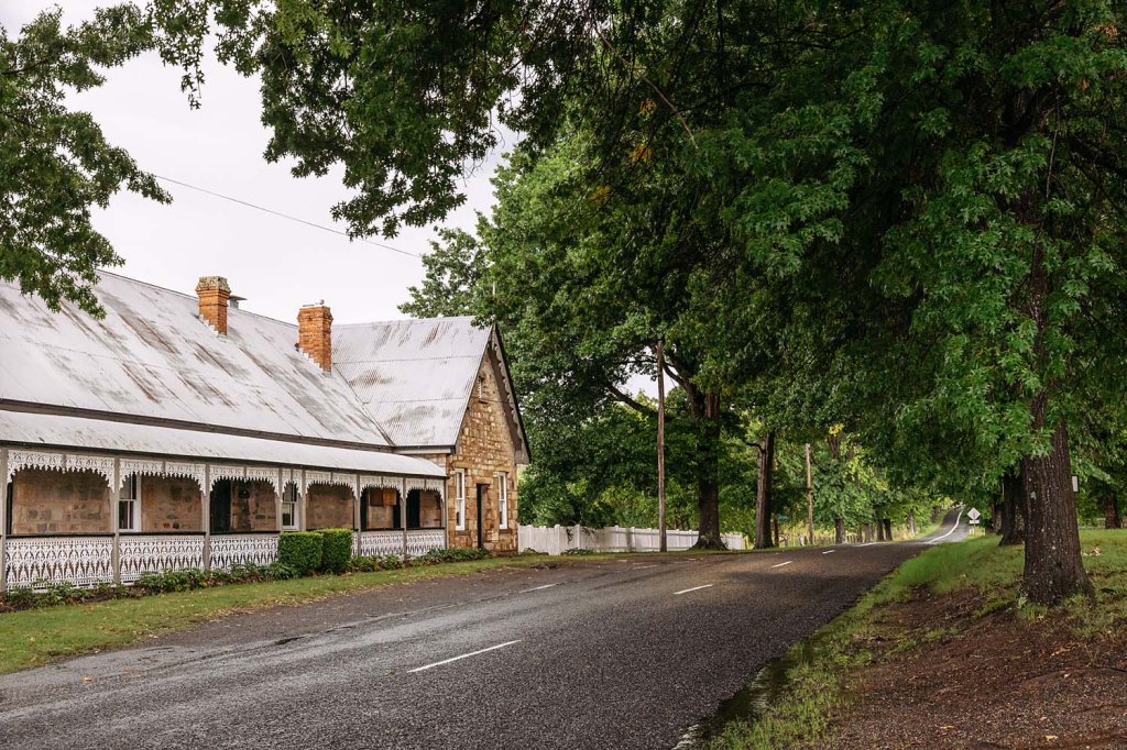 A quiet rural road lined with trees, featuring a stone house with a covered porch and a weathered roof.