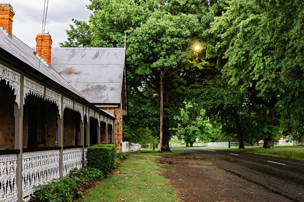A vintage building with a corrugated metal roof and ornate wooden porch railings, surrounded by lush greenery and a quiet road under a cloudy sky.